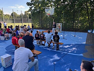 Kinder und Jugendliche sitzen auf Bänken und Sitzwürfeln auf dem Basketballplatz, Bürgermeister Seidl steht vor ihnen.